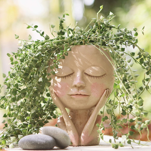a womanly face pot with closed eyes and arms on her cheeks with a trailing succulent flowing out of the head like hair